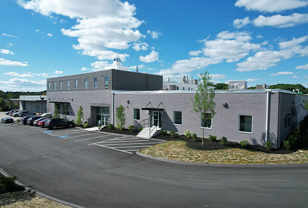 A modern, gray industrial building under a partly cloudy sky. It has a flat roof and several windows. In front, there are parked cars and a driveway. A small grassy area with young trees and shrubs surrounds the structure.