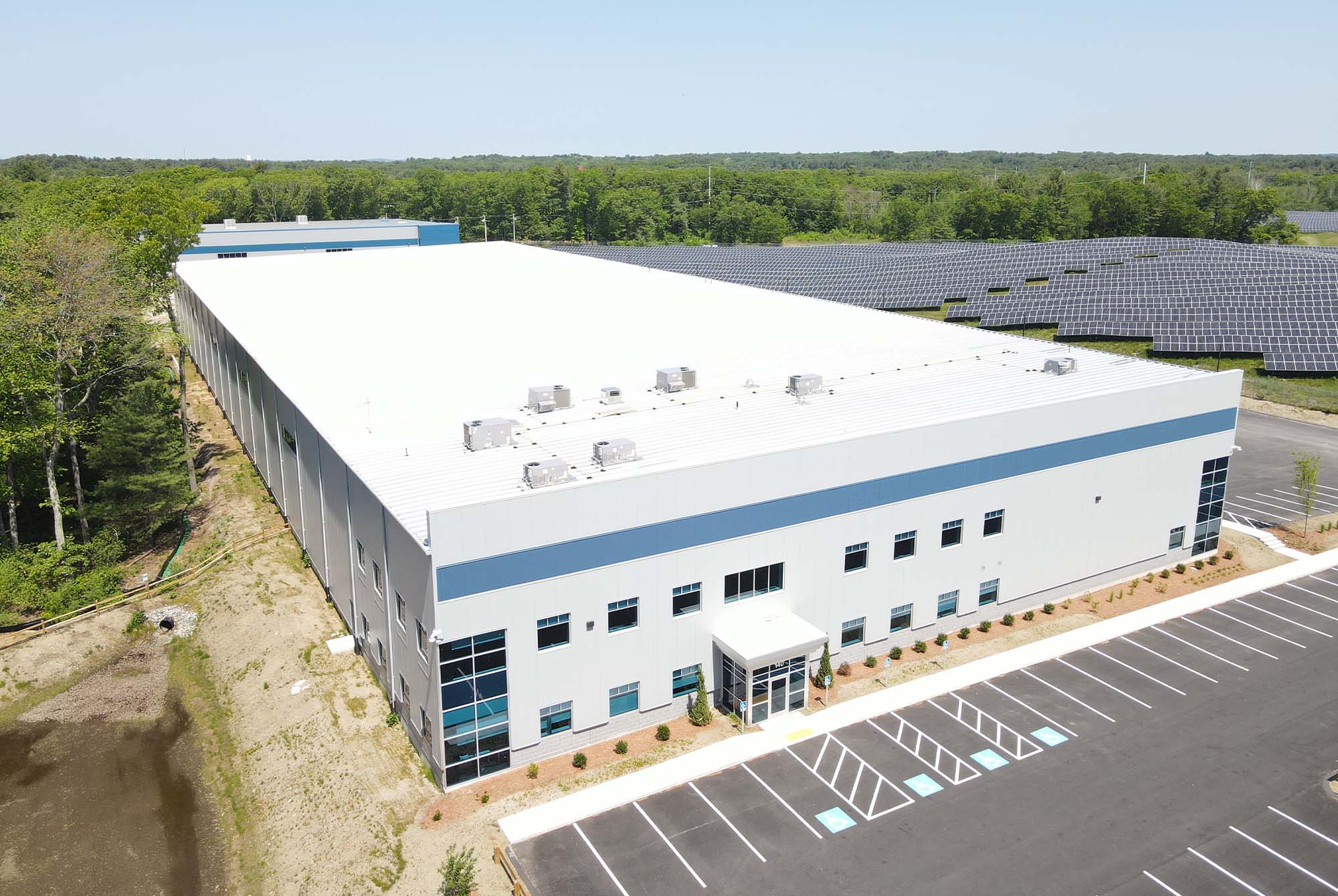 A large, rectangular industrial building with a white roof and gray walls, surrounded by a parking lot. Solar panels are visible in the background. Trees and greenery border the area under a clear blue sky.