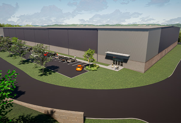 A modern industrial warehouse with a flat roof, surrounded by a parking area with several cars. The building is set on a green lawn with trees, and a paved road runs past the front. The sky above is partly cloudy.