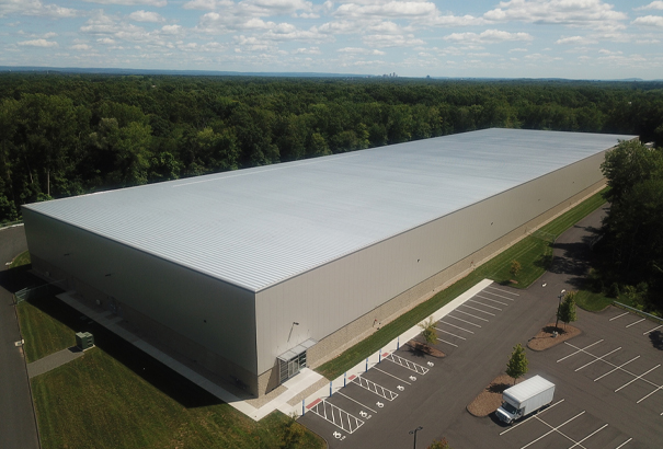 Aerial view of a large, rectangular warehouse with a light gray roof surrounded by a mostly empty parking lot, set in a green, wooded area. The sky is partly cloudy.