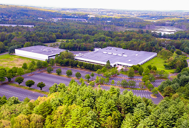 Aerial view of a large industrial building surrounded by a mostly empty parking lot. The area is lush with trees and greenery, and the horizon shows a mix of forested land and distant hills under a light sky.