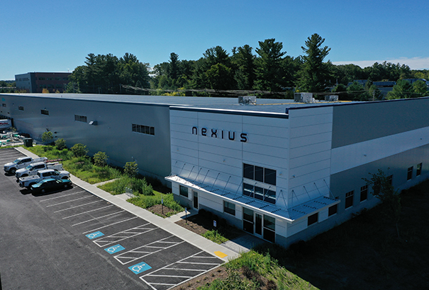 A large industrial building with Nexius on the facade, surrounded by trees. The parking lot in front has several parked cars and designated accessible parking spaces. The setting is sunny with a clear blue sky.