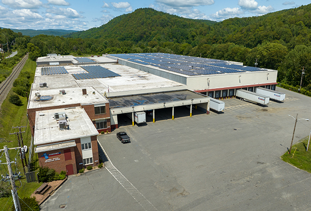 Aerial view of a large industrial warehouse with solar panels on the roof, surrounded by green trees and hills. The building has loading docks with several trucks parked nearby, and an empty asphalt parking lot in the foreground.