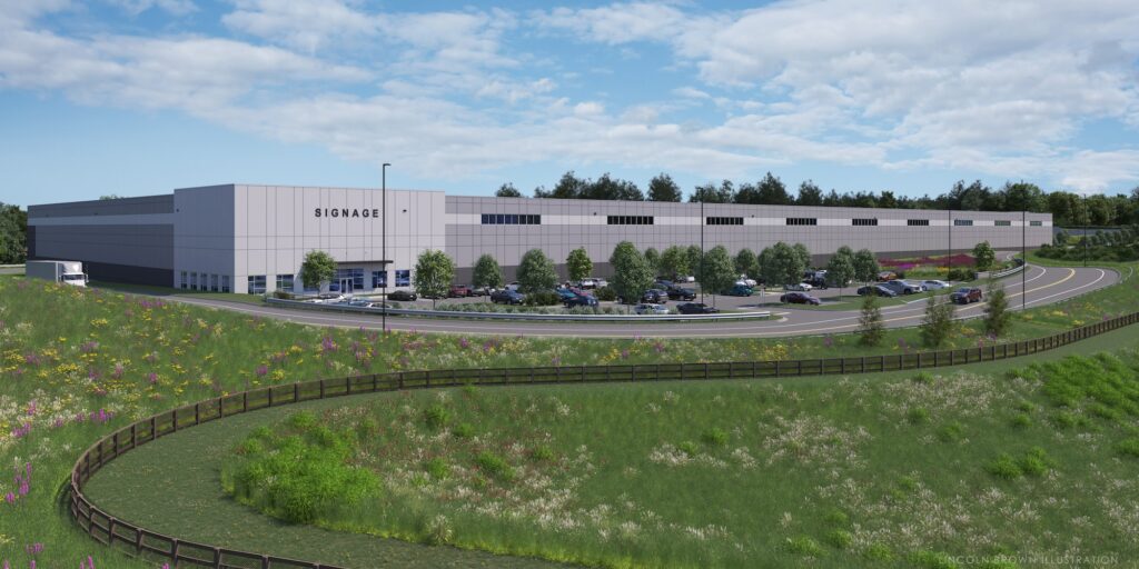 Large industrial warehouse with a sign reading SIGNAGE surrounded by parking lots and trees. A road curves in the foreground with a wooden fence and grassy fields. The background features forested areas and a partly cloudy sky.
