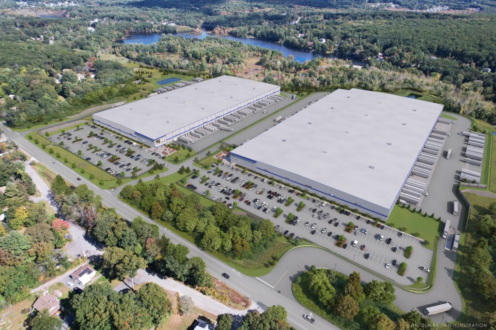 Aerial view of two large, adjacent warehouse buildings surrounded by parking lots filled with cars. Truck docks are visible on the buildings. The scene is set in a wooded area near a lake, with trees and roads around the complex.