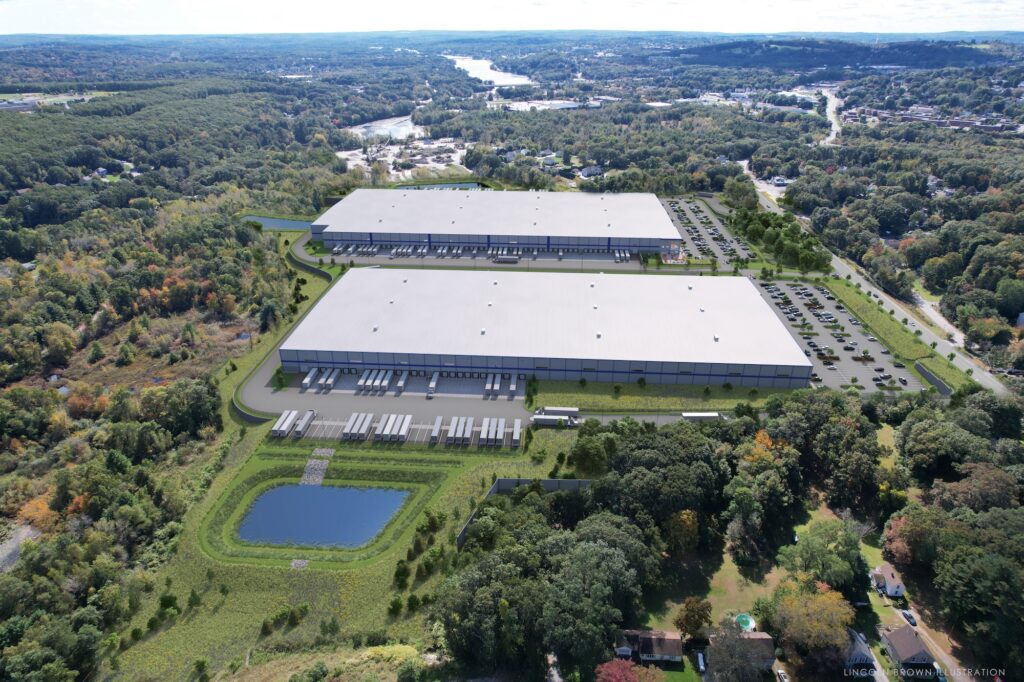 Aerial view of two large industrial warehouse buildings surrounded by parking lots, greenery, and a pond. In the distance, a river winds through a landscape of trees and scattered buildings under a cloudy blue sky.