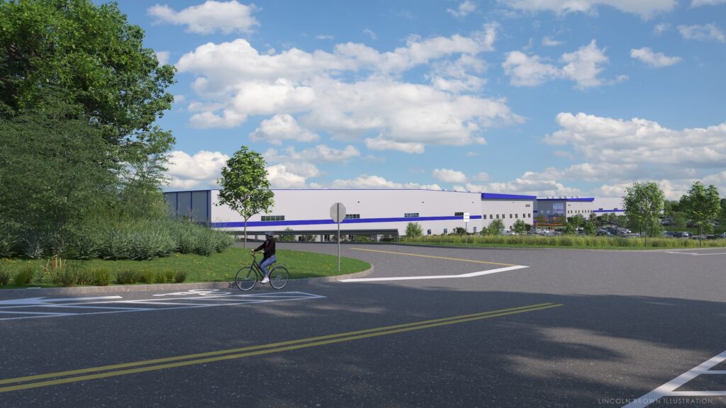 A cyclist rides on a bike path near a large warehouse building under a blue sky with scattered clouds. The building is surrounded by greenery and trees, and a road runs alongside it.