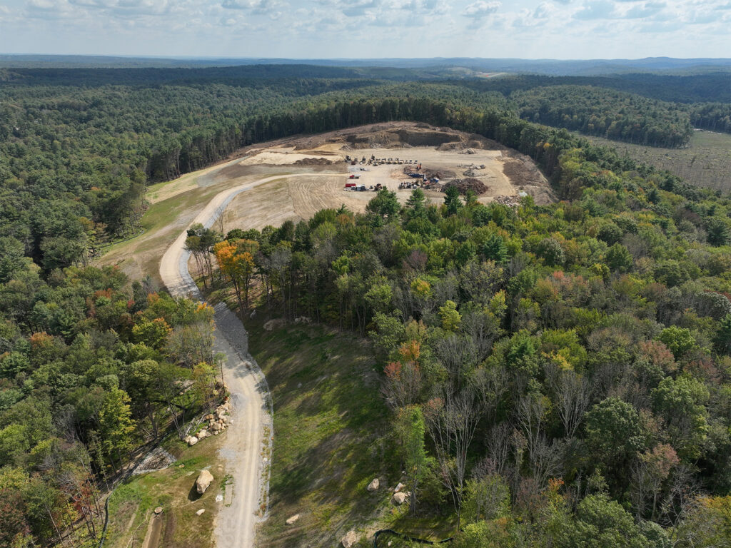 Aerial view of a winding dirt road leading to a large clearing in a dense forest. The clearing contains construction equipment and vehicles. Lush green trees surround the area with a distant horizon under a partly cloudy sky.