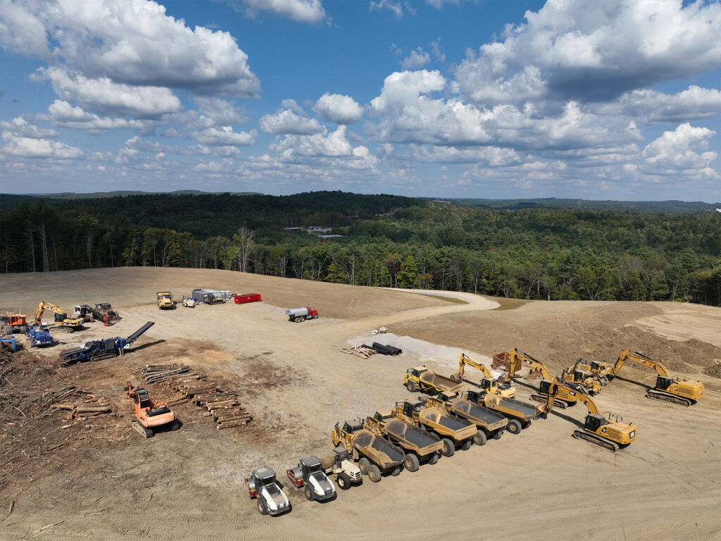 Aerial view of a construction site with various heavy machinery, including excavators and dump trucks, organized in rows. Logs are piled nearby, and a few vehicles are parked. The site is surrounded by dense green forest under a partly cloudy sky.
