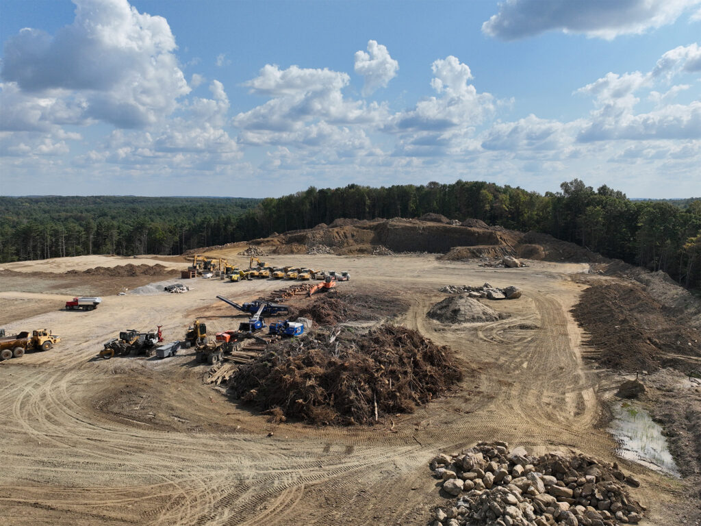 Aerial view of a construction site featuring various heavy machinery, including trucks and excavators. The landscape is dusty with piles of earth and debris. A dense forest borders the site in the background under a partly cloudy sky.