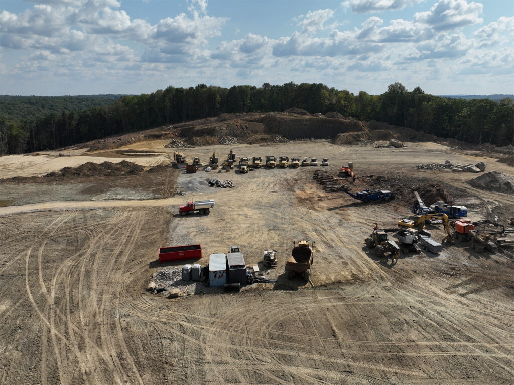 Aerial view of a construction site with various heavy machinery, vehicles, and equipment scattered on a dirt field. Piles of earth and trees border the area. The sky is partly cloudy, with a forest visible in the background.