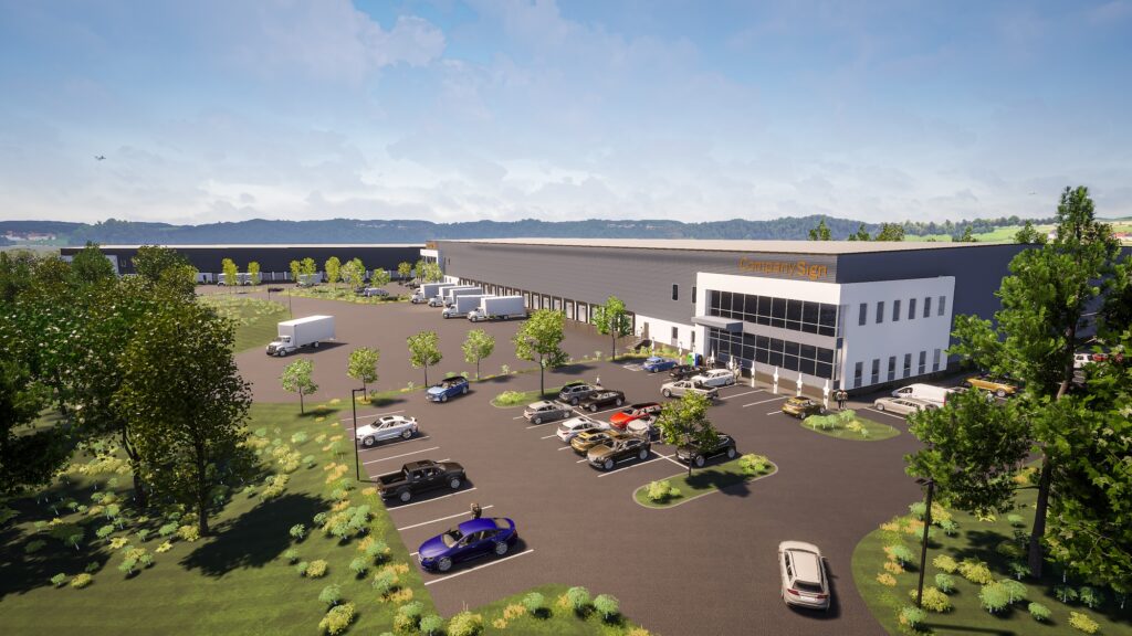 Aerial view of a large industrial warehouse with Kronoby S43 signage. The facility is surrounded by a parking lot with several cars, green trees, and open grassy areas. The background features distant hills under a partly cloudy sky.