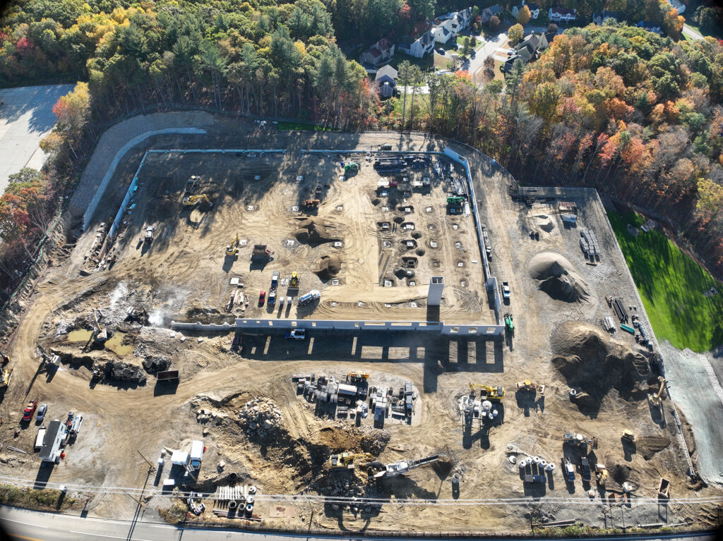 Aerial view of a construction site surrounded by trees and residential areas. The site features a dirt surface with machinery, vehicles, and materials, including concrete structures, mounds of earth, and equipment spread across the area.