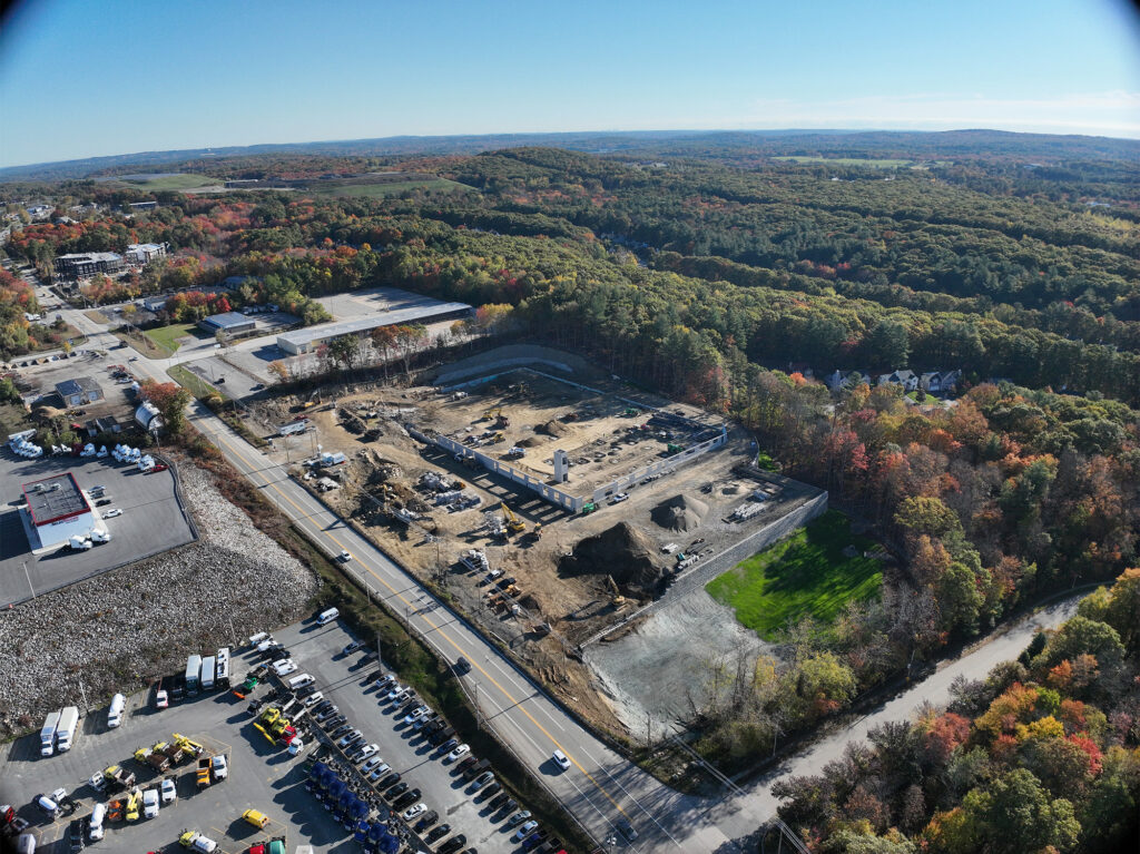 Aerial view of a construction site surrounded by dense, colorful autumn foliage. The site features various construction vehicles and materials. Nearby roads and parking areas are visible, along with several buildings and expansive forested land.