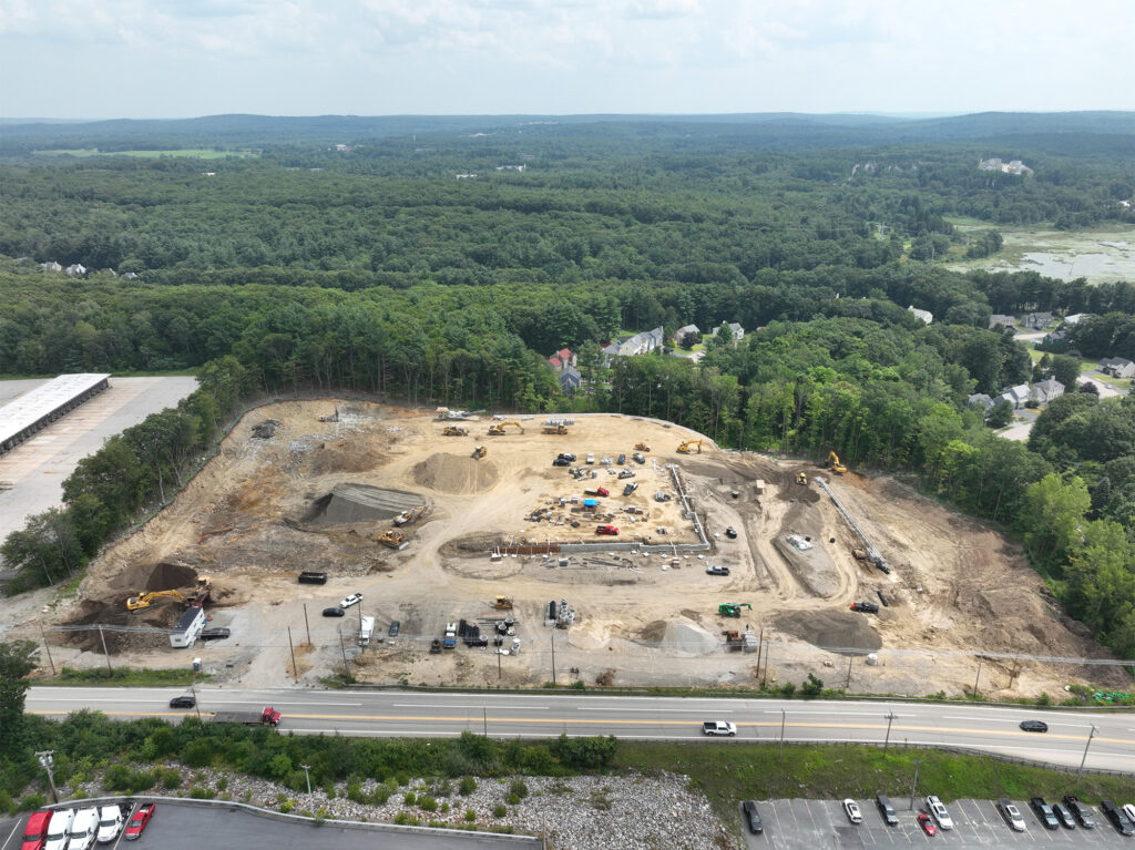 Aerial view of a construction site surrounded by trees. The site contains heavy machinery, vehicles, and dirt piles, with a road and parked cars in the foreground. The background shows a forested landscape under a cloudy sky.