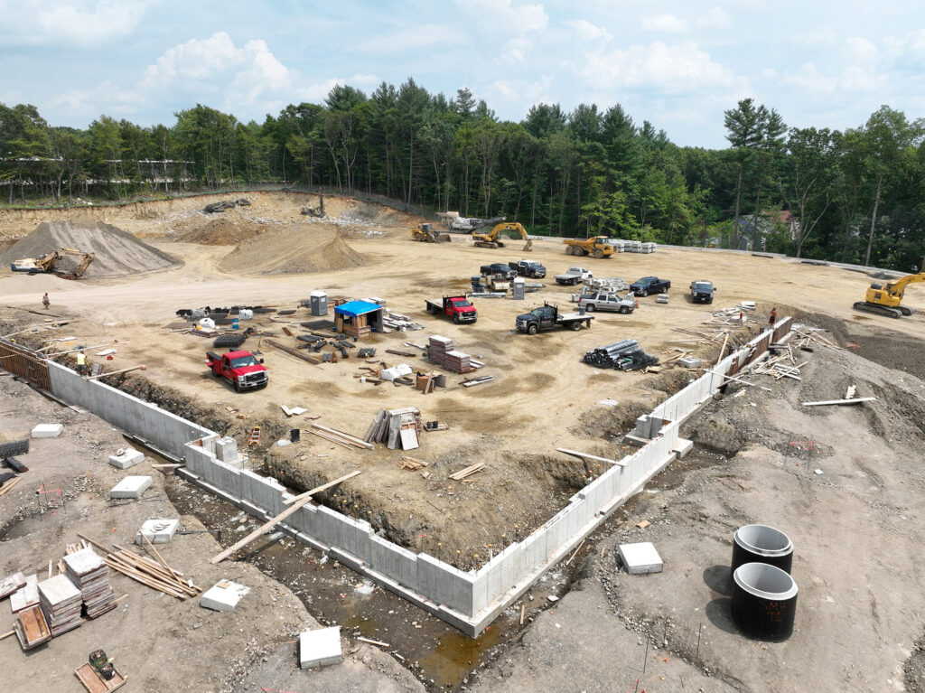 A construction site with a foundation being built in a clearing. Several trucks and construction equipment are scattered around. Piles of materials and debris are visible. Trees surround the site, and the sky is partly cloudy.