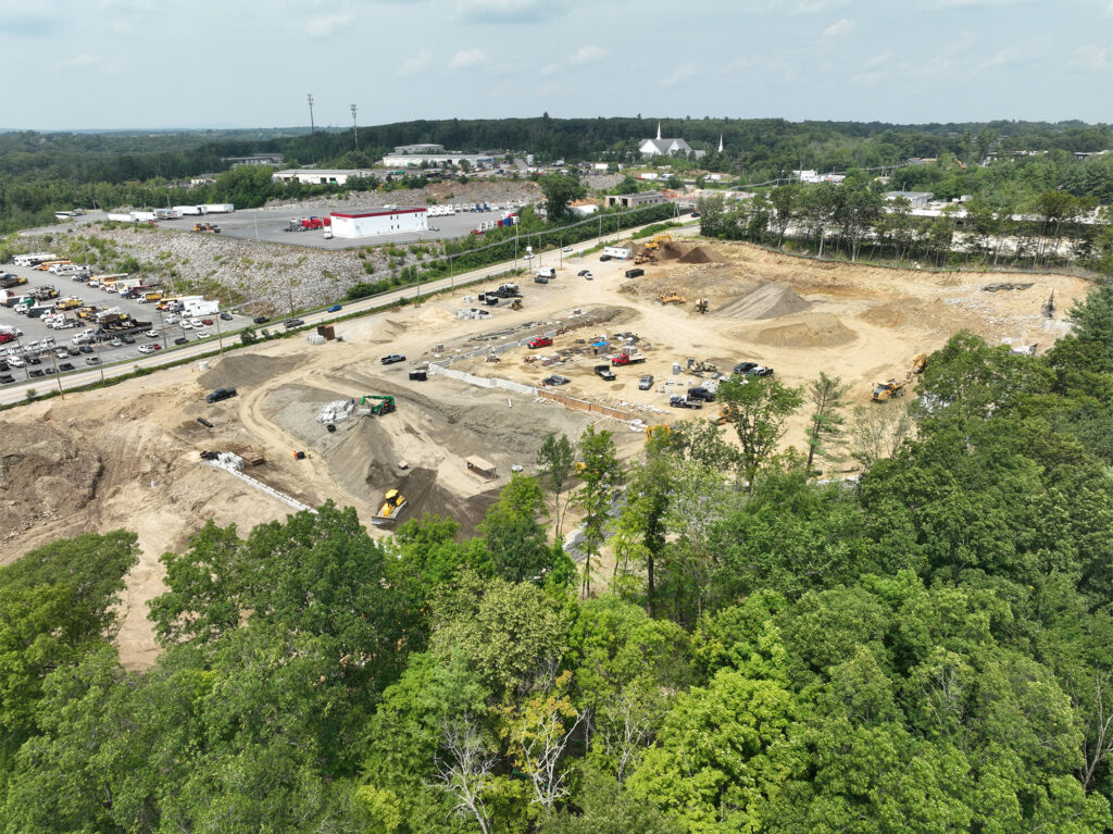 Aerial view of a large construction site with vehicles and equipment, surrounded by trees. Nearby are parking lots and industrial buildings. The sky is partly cloudy.
