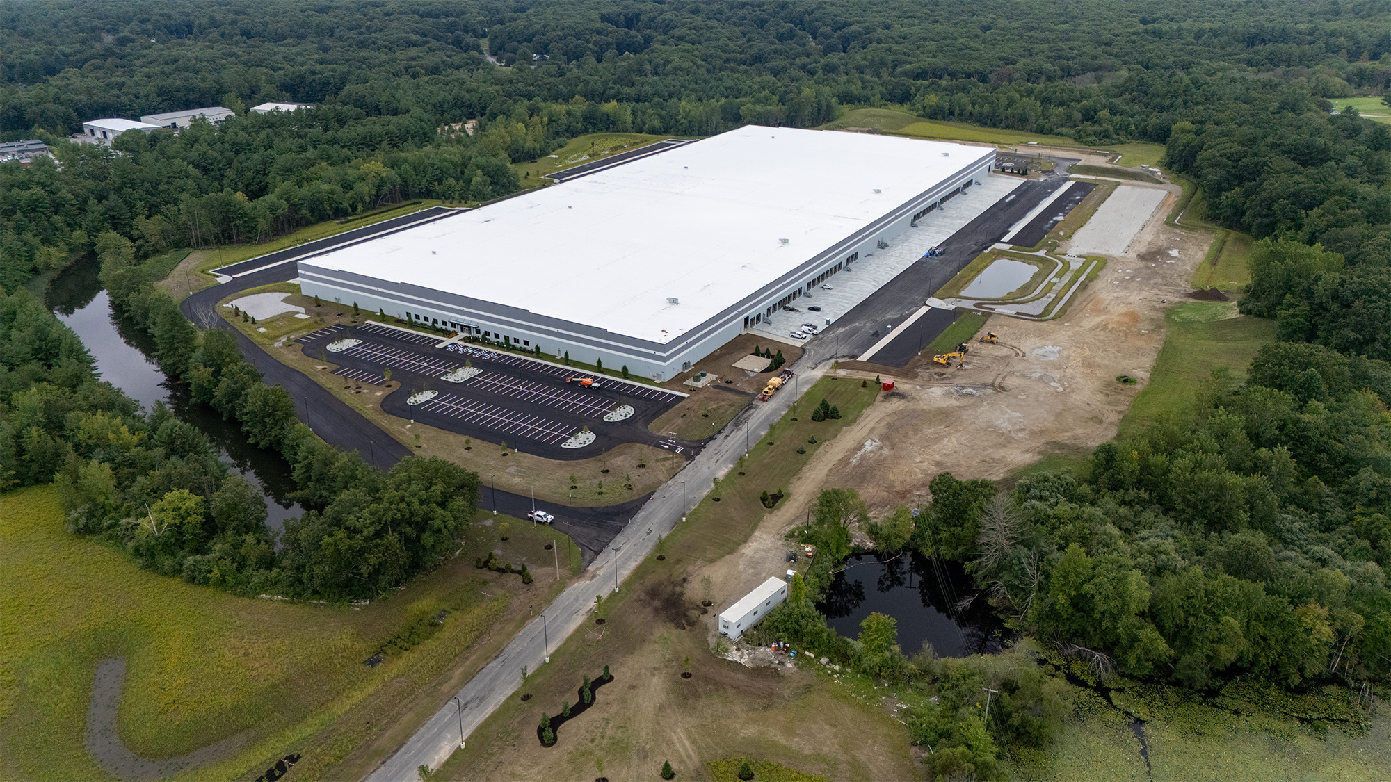 A large warehouse with a white roof surrounded by green forest. The building has multiple loading docks, a parking area, and paved roads. An adjacent pond and open spaces are visible in the landscape.