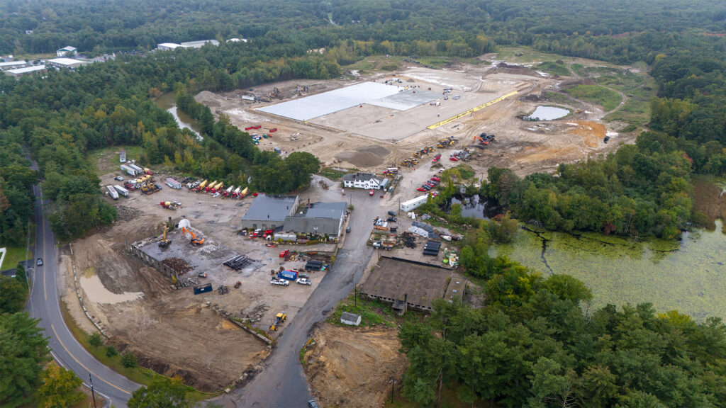 Aerial view of a construction site surrounded by dense woodland. The site features large areas of cleared land, construction vehicles, and partially built structures. Theres a road curving through the bottom left of the image and a pond to the right.