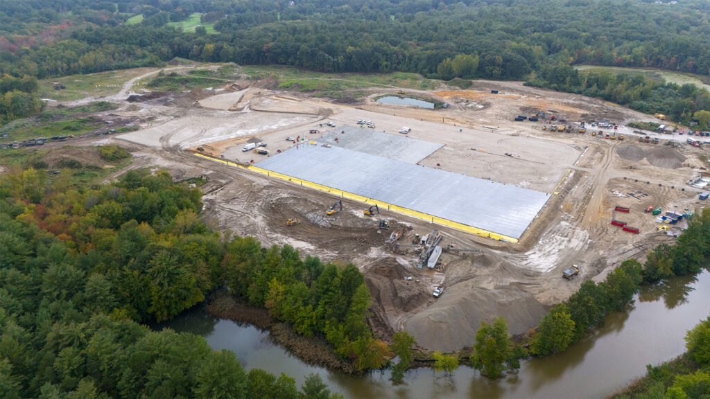 Aerial view of a large construction site surrounded by dense green forest. A massive, rectangular concrete slab is being developed, with construction vehicles and equipment scattered around the area. A river runs adjacent to the site.