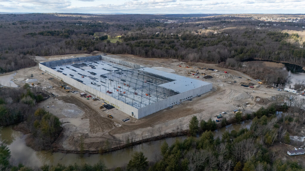 Aerial view of a large warehouse under construction in a clearing surrounded by forests. The building has partial walls and visible steel framework with construction vehicles around it. Sparse clouds and distant hills are in the background.