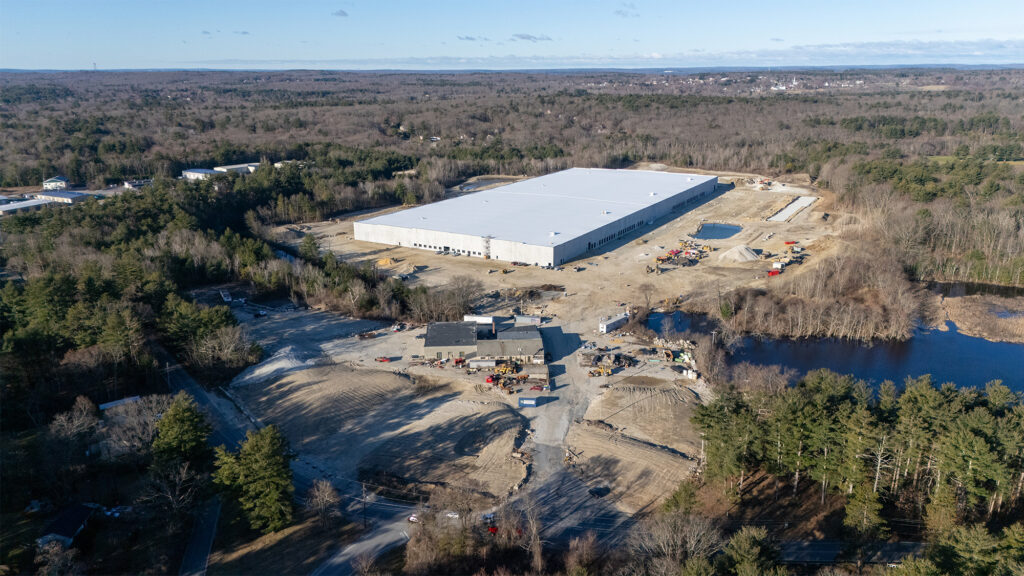 Aerial view of a large warehouse under construction surrounded by a forested area. Theres a partially cleared area with construction vehicles and equipment nearby. A small body of water is visible to the right. The sky is clear and the landscape is expansive.