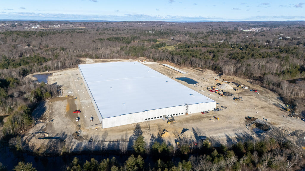 Aerial view of a large, white-roofed industrial building under construction surrounded by cleared land and forest. Construction vehicles and equipment are visible. The surrounding area is wooded with patches of open land.