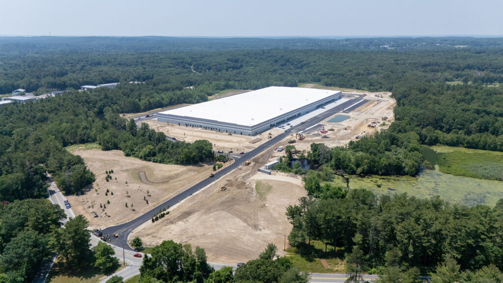 Aerial view of a large, newly constructed warehouse surrounded by trees. The building is rectangular with a white roof. Nearby, there are paved roads and construction vehicles. The background features a landscape of dense forest under a clear sky.