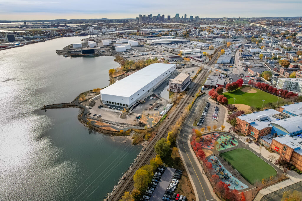 Aerial view of a waterfront industrial building, adjacent to a park with a green field and colorful playground, set near train tracks, with a city skyline visible in the distance under a cloudy sky.