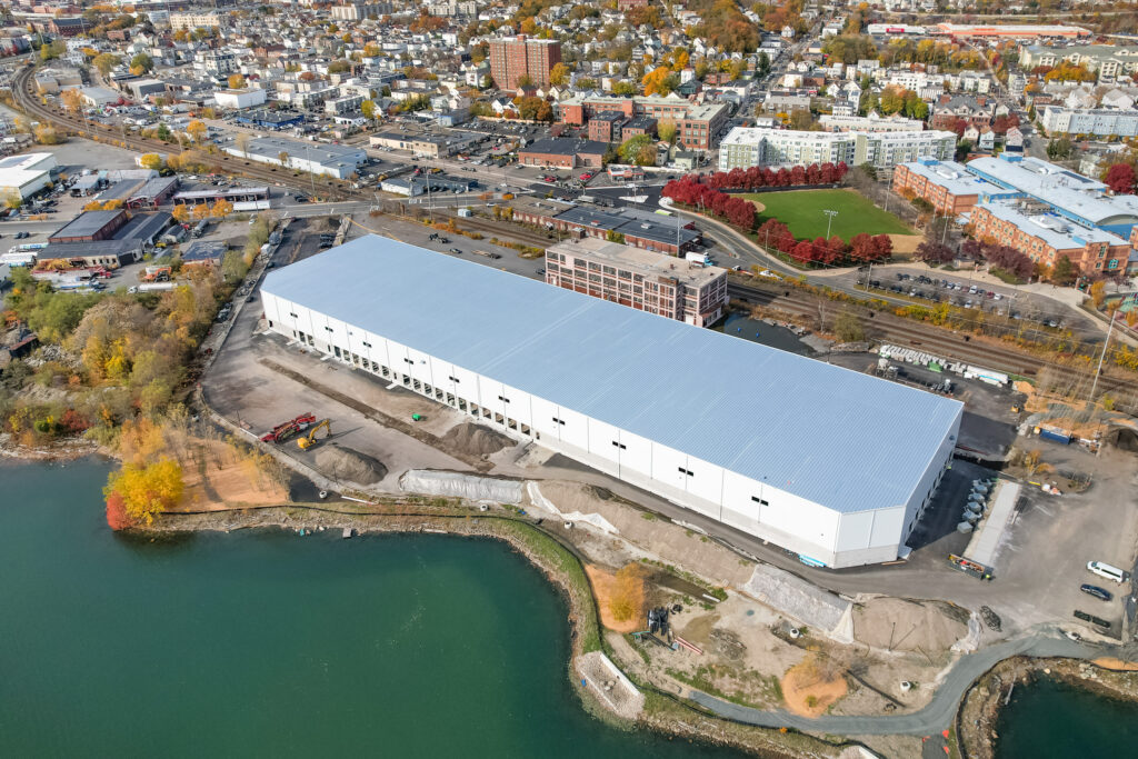 A large white industrial building sits near a waterfront, surrounded by roads, trees with autumn foliage, and a nearby city neighborhood in the background.