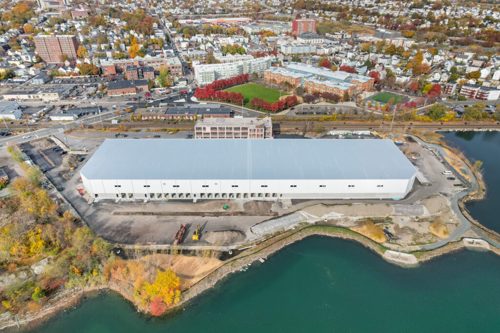 Aerial view of a large white warehouse by the water, surrounded by industrial lots, roads, and fall-colored trees, with a residential neighborhood and apartment buildings in the background.