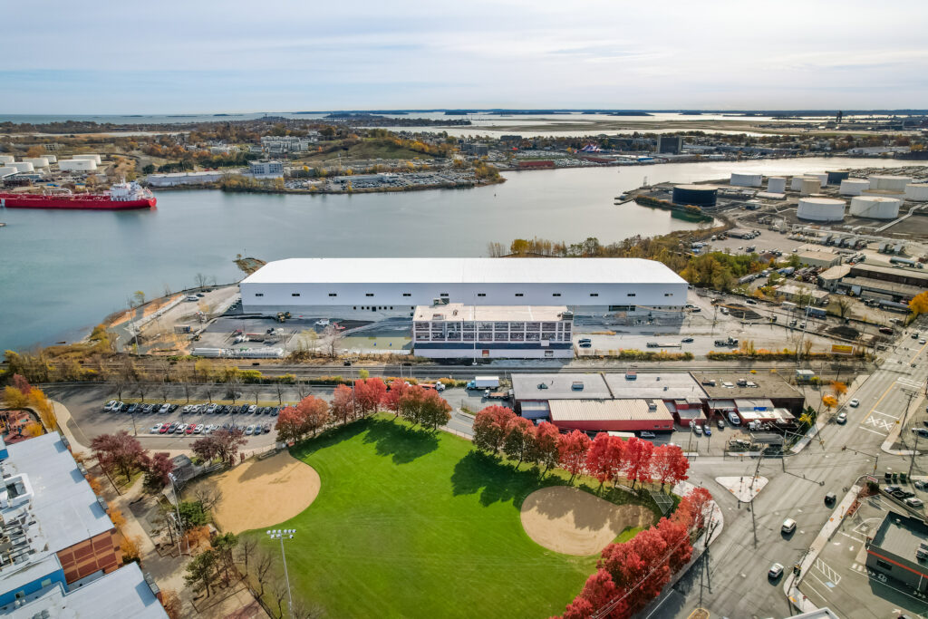 Aerial view of a waterfront industrial area with a large white warehouse, parking lots, a baseball field with green grass, red trees, and water in the background.