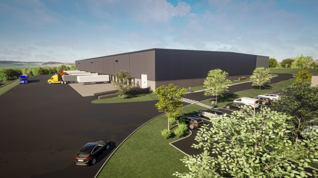 A large industrial warehouse with a gray exterior sits surrounded by a parking area filled with cars. Nearby, semi-trucks, including a blue and white one with trailers, are parked at loading docks. Trees and greenery line the expansive lot under a partly cloudy sky.