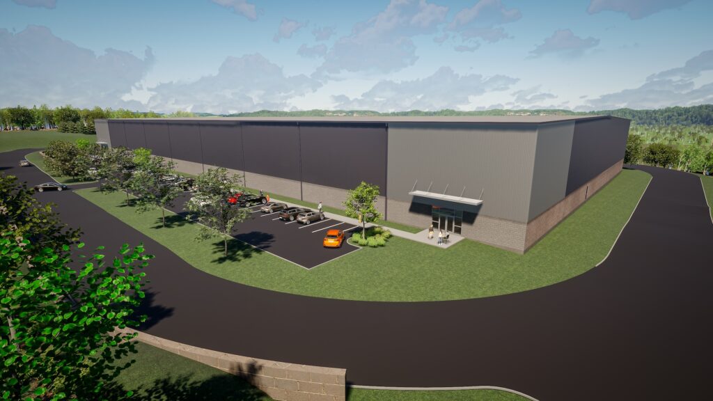 Aerial view of a large industrial warehouse with a gray facade and landscaped surroundings. Several cars are parked in the adjacent lot. A road encircles the building, and trees are dispersed throughout the green areas.