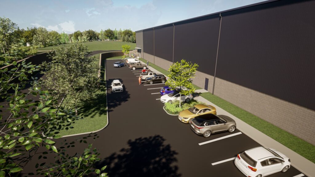 A long, dark gray warehouse is adjacent to a parking lot with several parked cars. Green lawns and trees surround the area, with a path leading towards a wooded area in the background. The sky is partly cloudy.