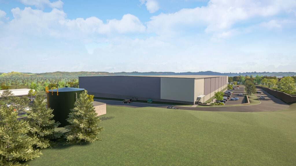 A large, modern warehouse building in a grassy area with a few trees in the foreground. Several cars are parked in the adjacent lot. The sky is partly cloudy.