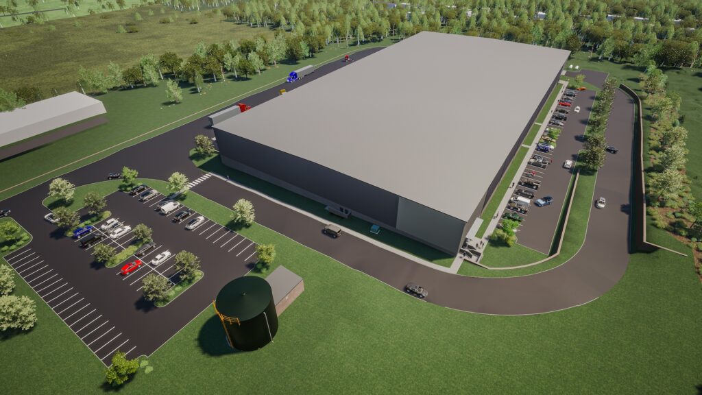 Aerial view of a large warehouse surrounded by parking lots with cars and trucks. The building is situated in a green area with trees and a road leading to the facility. The landscape includes grassy fields and additional structures nearby.