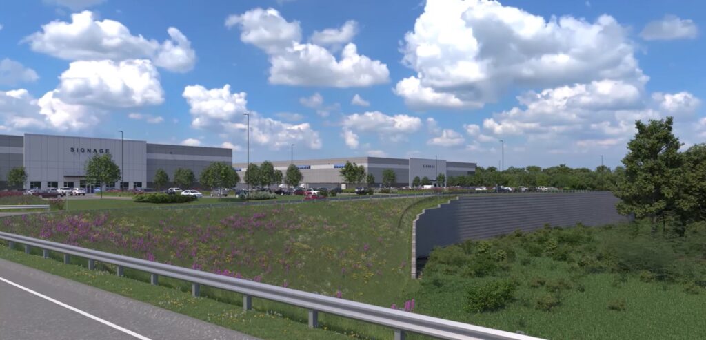 A large industrial building with several loading bays is surrounded by a grassy landscape and purple flowers. Multiple cars are parked in front. A road and guardrail are visible in the foreground under a cloudy blue sky.