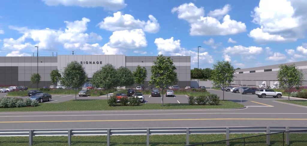 A parking lot with several cars is in front of a large industrial building. The building has SIGNAGE written on it. There are trees planted around the lot, and fluffy clouds are visible in the blue sky above.