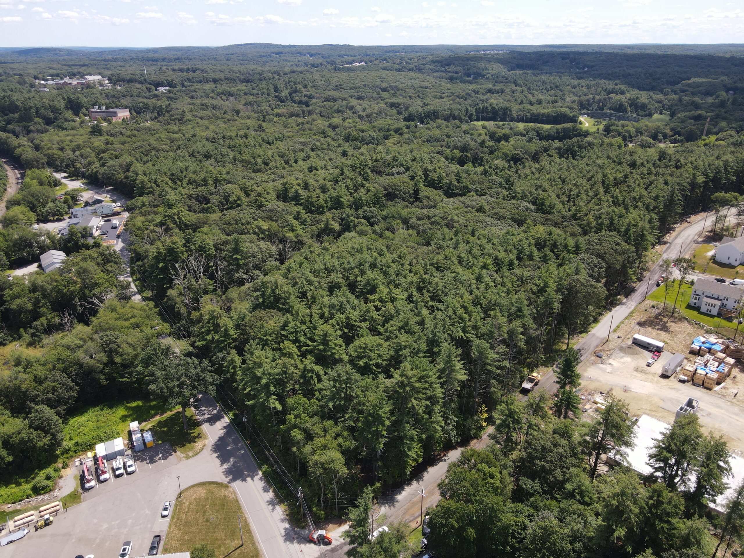 Aerial view of a vast, lush forest surrounded by a few roads and residential areas. Construction sites with scattered equipment are visible in the foreground. The sky is partly cloudy, extending over the expansive green landscape.