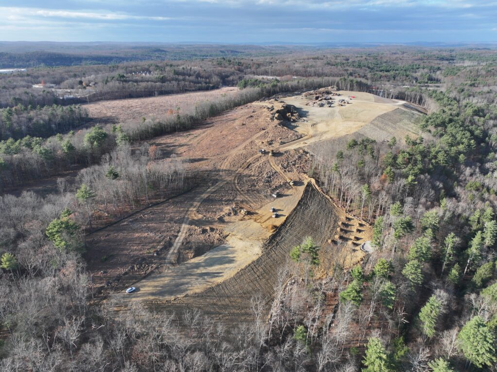 Aerial view of a large hillside construction site surrounded by forest. The area is cleared with visible soil and machinery. Various equipment and vehicles are seen on the cleared land amid patches of trees and a distant horizon.