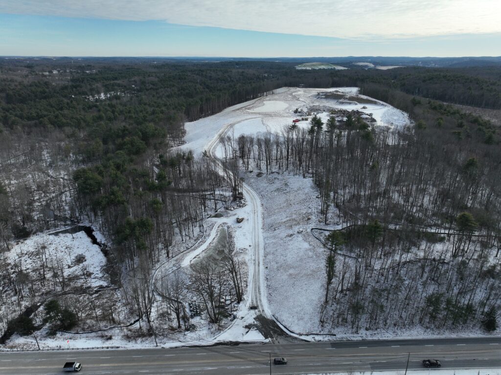 Aerial view of a snow-covered rural landscape with scattered trees and patches of bare ground. A winding road runs through the middle, with a highway in the foreground. Sparse vehicles are visible on the highway. The sky is overcast.