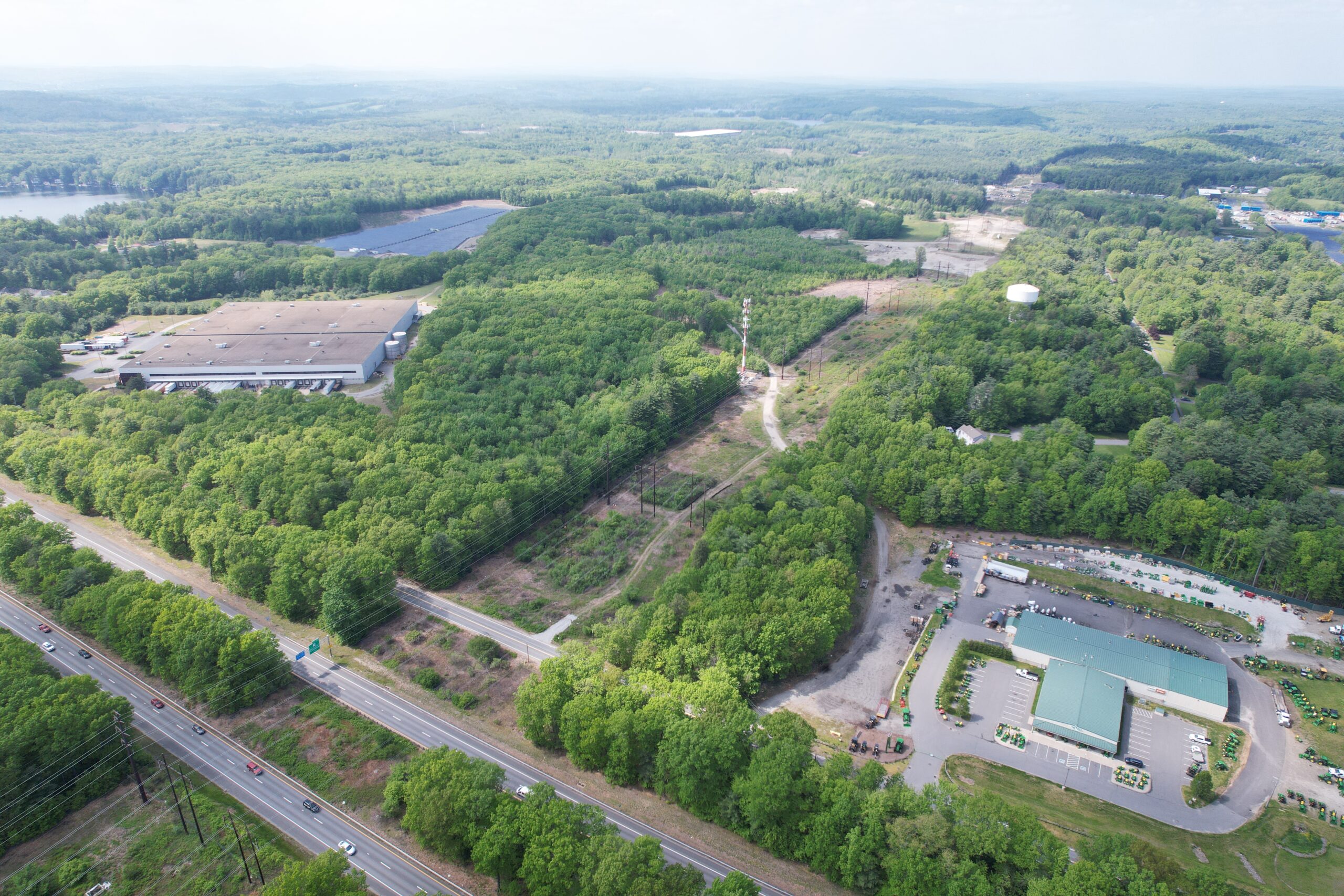 Aerial view of a green, forested area with a winding road. Theres a large building among the trees on the left and several smaller buildings with parked cars on the right. Power lines parallel a road in the lower part of the image.