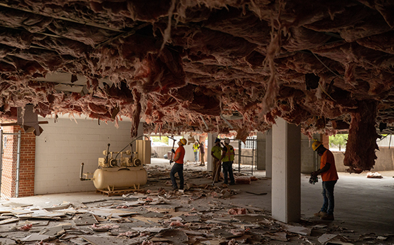 Workers in hard hats and safety vests clear debris in a building under construction or demolition. The ceiling is exposed with hanging insulation, and scattered materials cover the floor. Equipment is visible in the background.