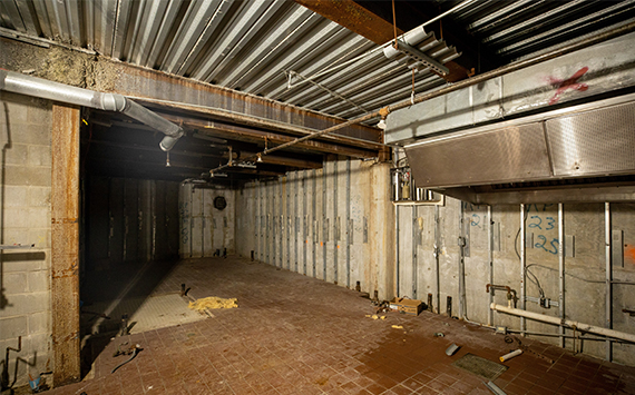 Dimly lit industrial room with exposed pipes, metal beams, and ventilation system. Brick floor shows signs of wear with scattered debris. Concrete walls have visible markings and rust. Overall appearance is aged and utilitarian.