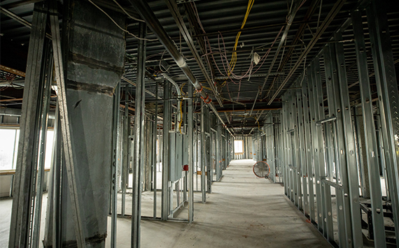 An unfinished building interior with exposed metal studs and framework. Wiring hangs from the ceiling, and natural light enters through large windows. The floor is bare concrete, and construction materials are scattered throughout.