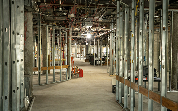 A large indoor construction site featuring metal framing for walls. Exposed wiring hangs from the ceiling, and various construction materials and tools are scattered around. Bright lighting illuminates the space.