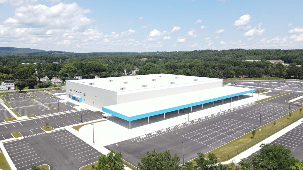 A large, modern white warehouse with a blue trim sits amidst empty parking lots on a clear day. The building is surrounded by green trees and hills in the distant background. The sky is blue with scattered clouds.