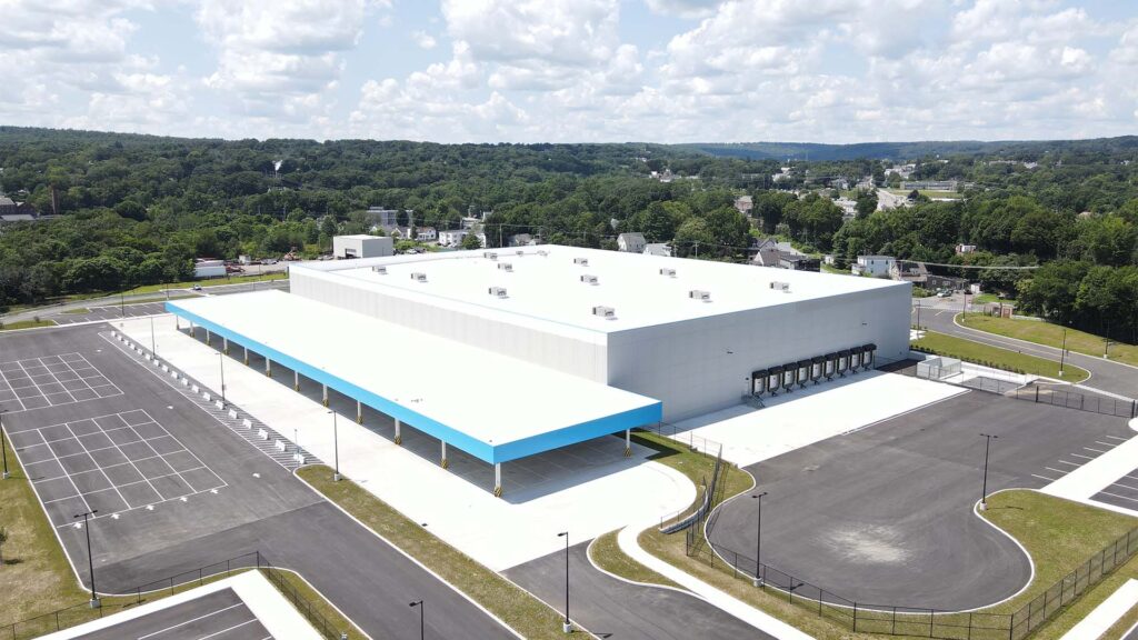 Aerial view of a large, modern warehouse with a white and blue facade, surrounded by an expansive parking lot. The building is set in a lush, green area with hills and forests visible in the distance under a partly cloudy sky.