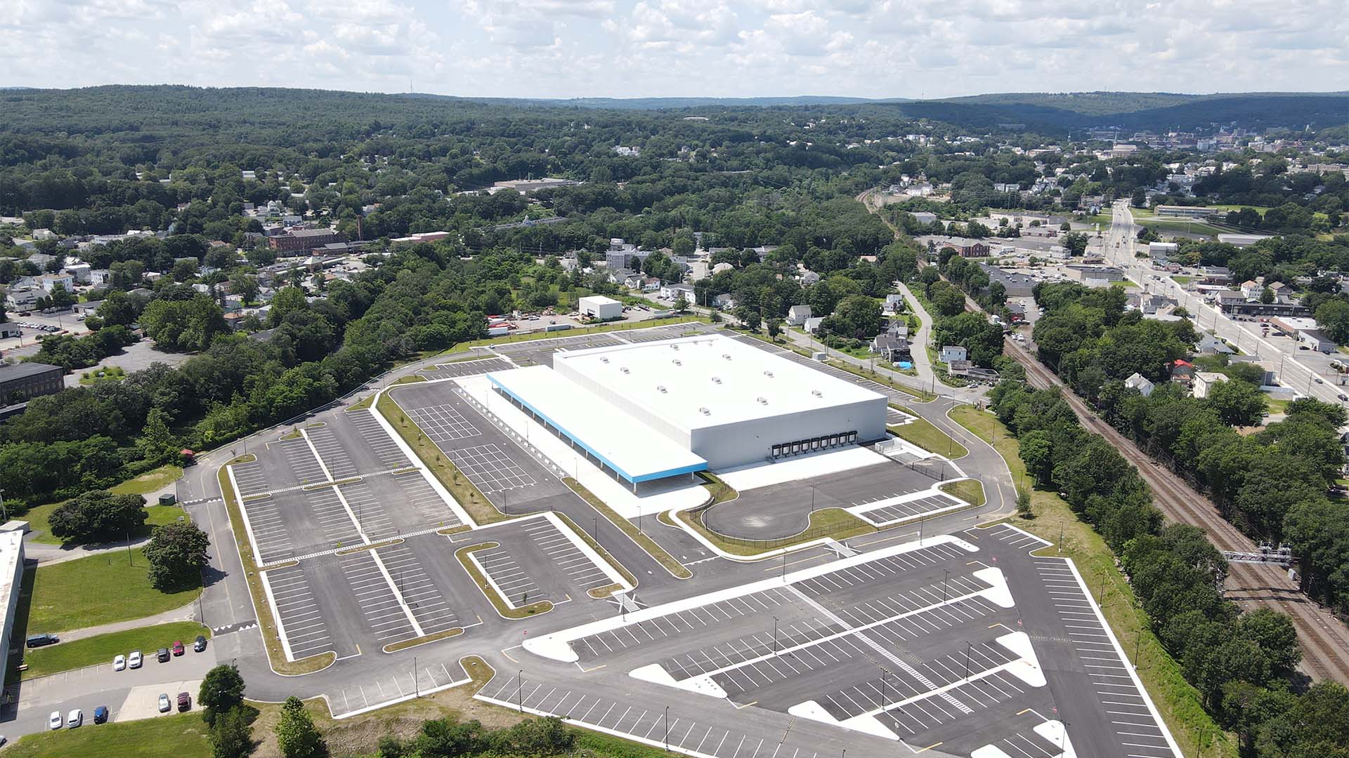 Aerial view of a large warehouse with a white roof, surrounded by mostly empty parking lots. The building is situated in a semi-urban area with trees and scattered buildings, under a partly cloudy sky.